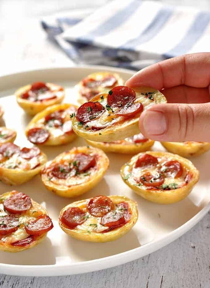Close up of a hand holding a Mini Pizza Potato Skin with a platter of Mini Pizza Potato Skins in the background.
