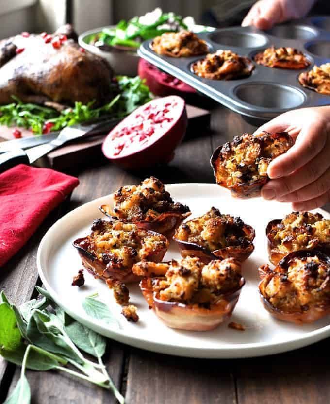 Hand reaching to take a Pork Sausage Apple Stuffing in Pancetta Cups (Muffin Tin) off a white plate.