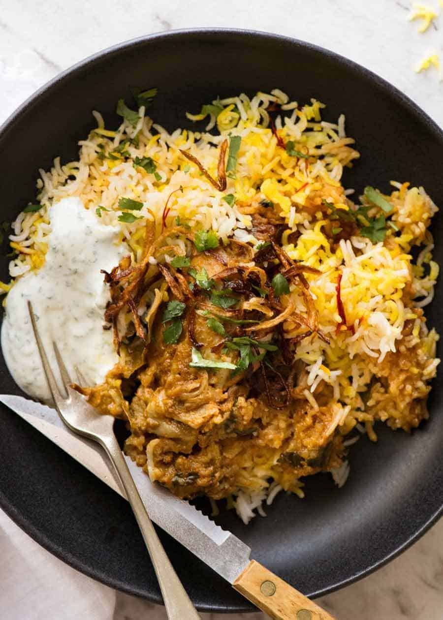 Chicken Biryani in a rustic black bowl with yellow saffron rice, garnished with crispy fried onions, coriander and minted yoghurt, ready to be eaten