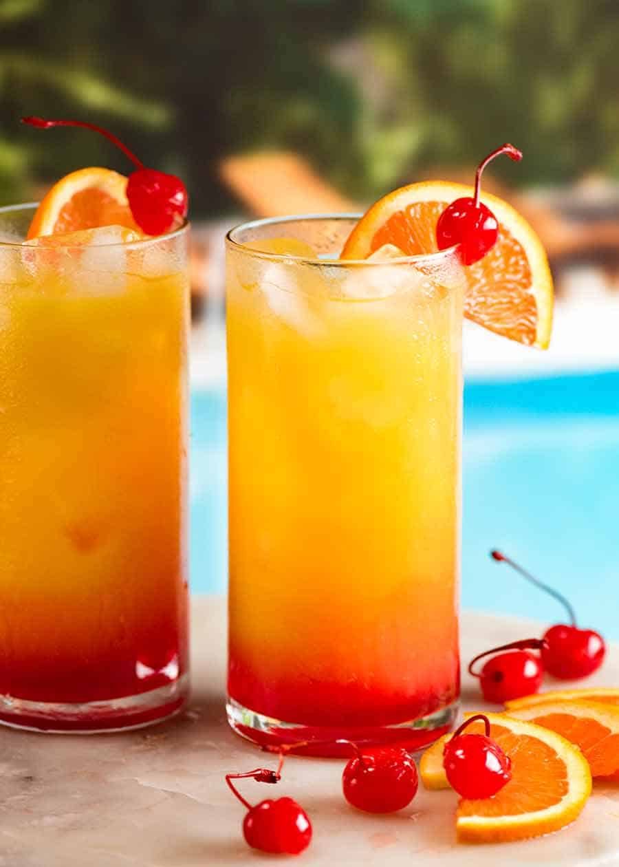 Two Tequila Sunrise cocktails by a pool, garnished with orange slice and maraschino cherries ready to drink